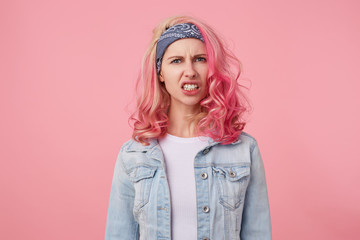 Outraged girl with pink hair shows her displeasure by baring her teeth, looks displeased at the camera, stands over pink background.