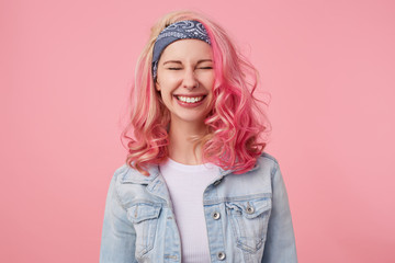 Happy cute smiling lady with pink hair and tattooed hands, waiting for surprise with closed eyes, broadly smiling, standing over pink background, wearing a white t-shirt and denim jacket.