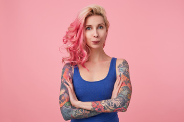 Portrait of funny cute lady with pink hair and tattooed hands, standing over pink background and looking at the camera, wearing a blue shirt. People and emotion concept.