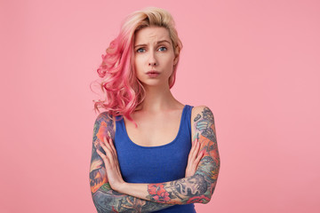 Young sad beauty woman with pink hair, stands with crossed arms over pink background, looks displeased and unhappy, wears a blue shirt. People and emoyion concept.