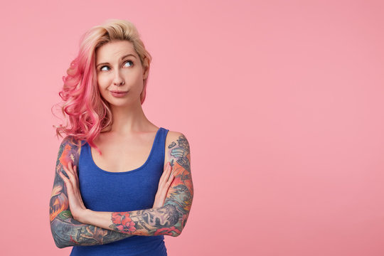 Portrait of young thinking cute lady with pink hair, wears blue shirt, stands over pink background copy space with crossed arms, looks up thoughtfully. People and emoyion concept.