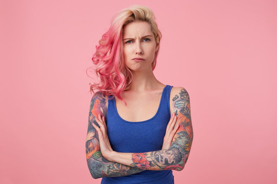 Young sad frowning beauty woman with pink hair, stands with crossed arms over pink background, looks displeased, wears a blue shirt. People and emotion concept.