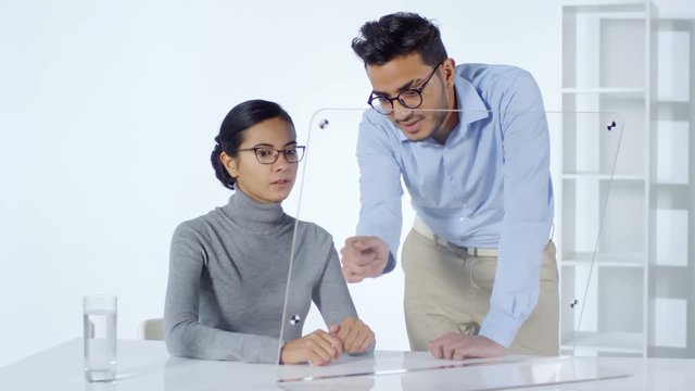 Lockdown of Asian businesswoman tapping and swiping on invisible interactive screen and talking to Arab male colleague standing nearby. Suitable for adding AR graphics