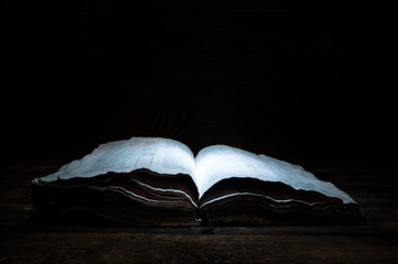 An old open book is lying on a wooden table in the dark. A light shines on the book from above....