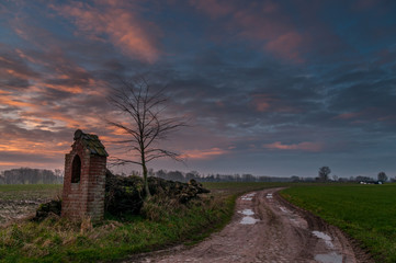 Impression of a worn-down memorial in the flemish country side, right next to an unpaved country road, flooded with pink light from an early morning sunrise.