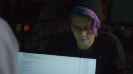 Medium shot of two young Caucasian hipsters sitting at computers in front of each other and talking about program code