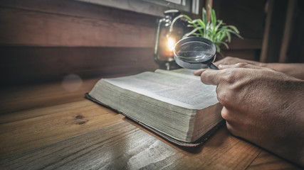 Close up hands of man searching text in Bible with magnifying glass on wood table with light of oil lamp.