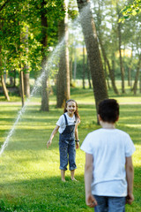Fototapeta premium Cheerful little girl and boy playing near watering on green grass and smiling. Brother and sister in wet clothes in the summer park. Best friends. Childhood concept.