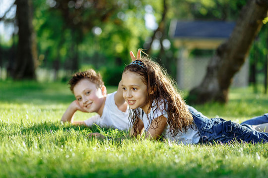 Cheerful little girl and boy laying down on green grass and smiling. Brother and sister in wet clothes in the summer park. Best friends. Childhood concept.