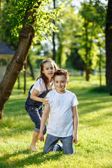 Fototapeta premium Cheerful little girl and boy play on green grass and smiling. Brother and sister in wet clothes in the summer park. Best friends. Childhood concept.