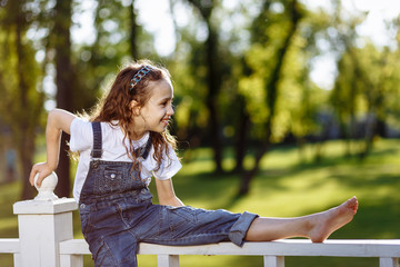 Naklejka premium Cheerful little girl sits on fence and smile in the summer park. Childhood concept.
