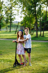 Fototapeta premium Cheerful little girl and boy drive the scooter on green grass. Brother and sister playing in the summer park. Best friends. Childhood concept.