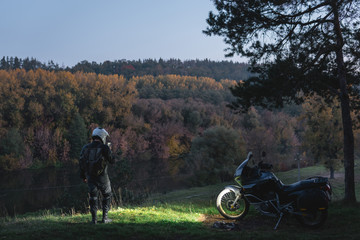 Rider standing with adventure motorcycle, Motorcyclist, A motorbike driver looks, headlights on, night forest, nature landscape. enduro travel road trip. Evening, autumn. Tourist traveler. copy space