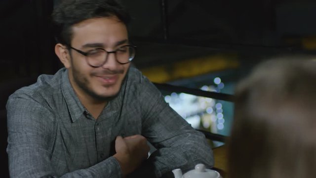 Medium shot of young Arab man sitting at caf&eacute; table and having warm talk to interlocutor sitting in front of him
