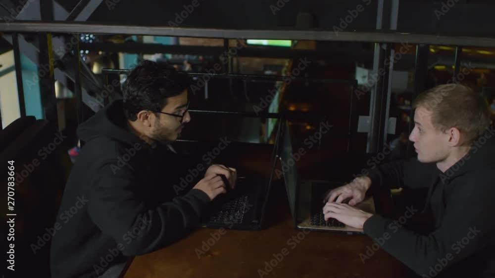 Poster tracking right of two young multiethnic men in hoodie sitting at laptops in front of each other and 