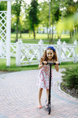 Cheerful little girl in rose dress drive the scooter near the wooden fence in summer park. Childhood concept.