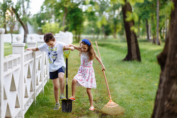 Cheerful little girl with rake and boy digging with shovel near the wooden fence in the park. Brother and sister works in garden. Childhood concept.