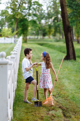 Fototapeta premium Cheerful little girl with rake and boy digging with shovel near the wooden fence in the park. Brother and sister works in garden. Childhood concept.