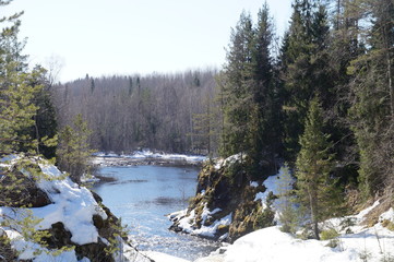 It's spring. On the banks of the river has not melted snow. The banks are rocky, the rocks grow pine and spruce. The day is clear, Sunny and cloudless. The water in the river is blue.