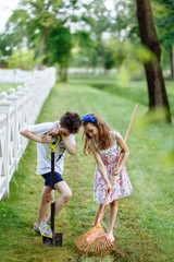 Fototapeta premium Cheerful little girl with rake and boy digging with shovel near the wooden fence in the park. Brother and sister works in garden. Childhood concept.