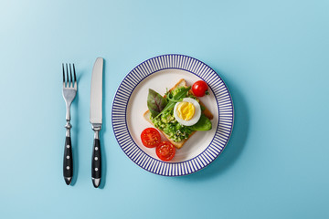 top view of toast with guacamole, boiled egg cherry tomatoes, fork and knife on blue background