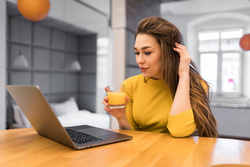 Obraz premium Beautiful young woman relaxing with her laptop while holding a glass of orange juice in the kitchen