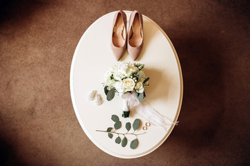 Brides wedding shoes with a bouquet with roses and other flowers on tha arm chair