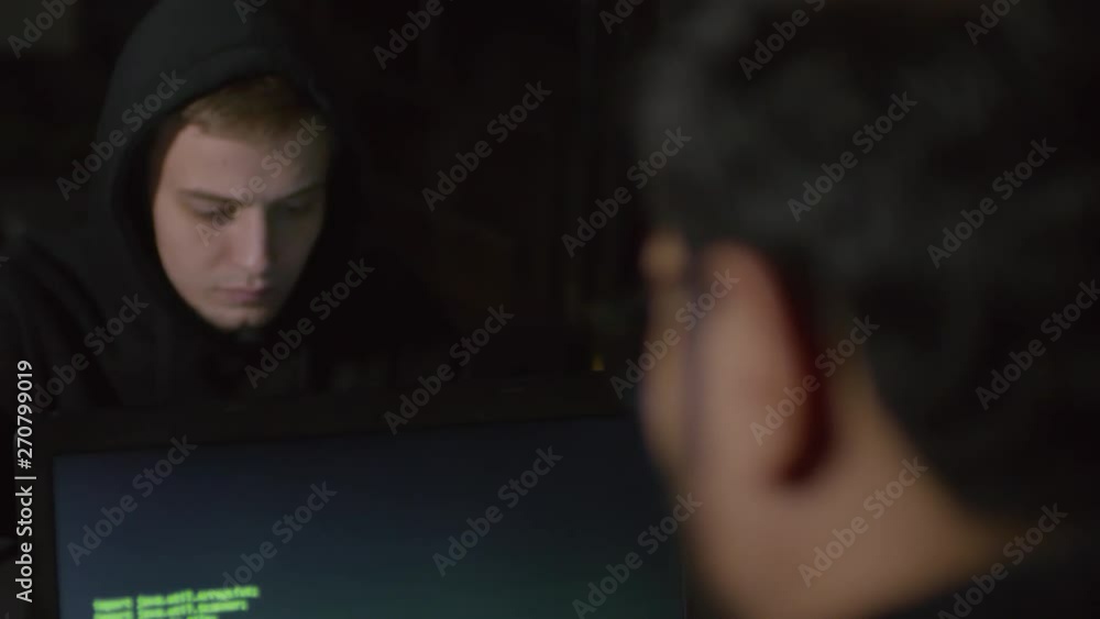 Poster rare view of two male hackers sitting at table in darkness, typing and getting access to databank