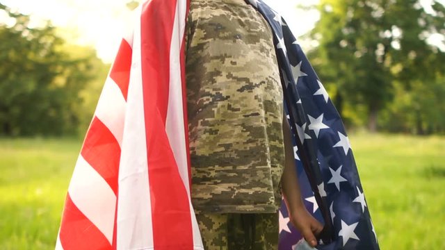 Curly boy in camouflage uniform stands covered by the American flag and smiles. Bottom-up view. Independence day usa
