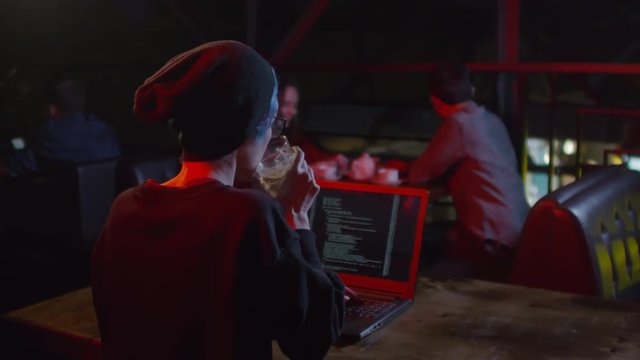 Back view of Caucasian female software developer sitting at laptop in caf&eacute;, typing program code, drinking cocktail while people on background having meal