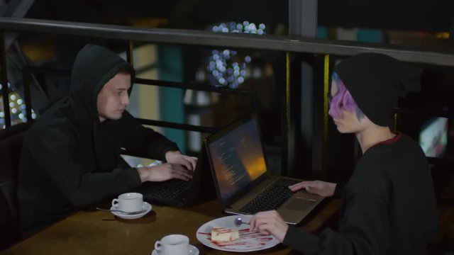 Lockdown of two hackers sitting at caf&eacute; table in front of each other and programming. Woman eating dessert and man drinking tea