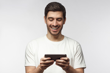Handsome laughing young man in white t-shirt, holding smartphone, playing video game, isolated on gray background