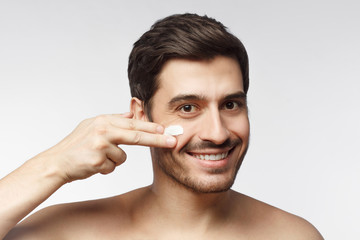Smiling shirtless young man applying facial cream, isolated on gray background. Skin care concept