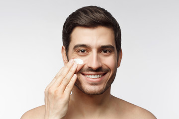 Close-up portrait of smiling shirtless young man applying facial cream, isolated on gray background. Skin care concept
