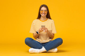 Happy young woman sitting on the floor, holding smartphone in hands and looking at camera, isolated...