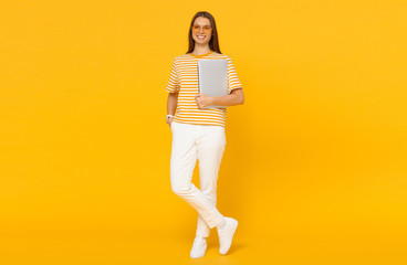Young woman in casual clothes holding laptop, isolated on yellow background
