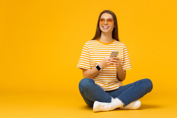 Happy young girl sitting on the floor, holding smartphone in hands and looking away, isolated on yellow background with copy space - Powered by Adobe