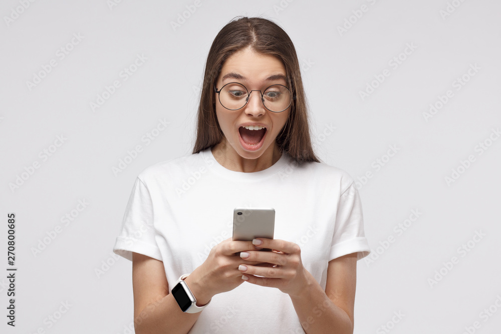 Wall mural close-up portrait of young female in white t-shirt, holding smartphone, looking surprised and shocke