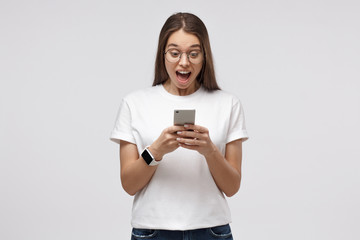 Fototapeta premium Portrait of young woman in white t-shirt, holding smart phone, looking schocked and surprised, isolated on gray
