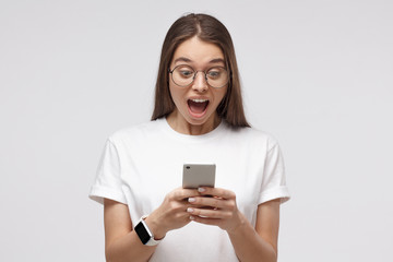 Close-up portrait of young female in white t-shirt, holding smartphone, looking surprised and shocked, isolated on gray background