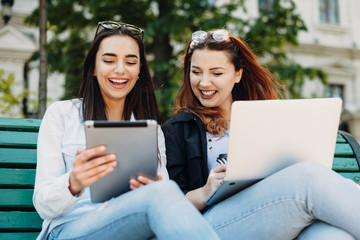Two lovely woman laughing while looking at a tablet screen while one is holding a laptop on the legs sitting on bench outside.