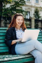 Portrait of a charming plus size woman sitting outside on a bench looking at her laptop on the legs smiling.