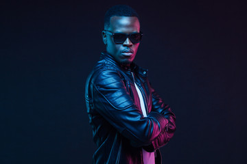 Studio neon portrait of stylish black man standing with crossed arms, wearing leather jacket and...