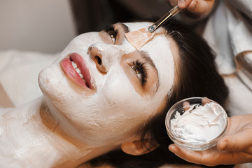 Close up portrait of a charming woman doing a white face mask in a wellnes spa. - obrazy, fototapety, plakaty