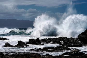 The angry sea, rough waters with a setting sun behind ominous clouds, emitting rays of light through the mist onto the angry sea, crashing wave upon the rocky shore.