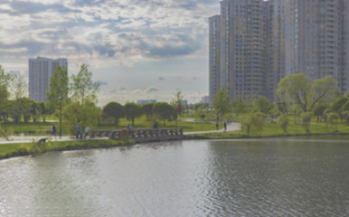 Fototapeta na wymiar Background. Blurred cityscape in the daytime. A pond, a pedestrian bridge, a park with people walking along the alleys, residential high-rise buildings against the sky with clouds.