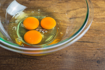 Three raw chicken egg yolk and white in glass bowl on top of wooden table top.