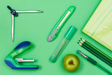 Book, color pencils, pen, felt-tip pen, paper knife, compass and stapler on green background.
