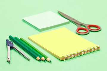 Compass, color pencils, notebook, note paper and scissors on green background.