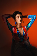 Vertical view of a woman in colorful clothes, seated on chair with closed eyes, isolated on a orange-coral background.